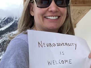 Maureen holds a handwritten sign reading "Neurodiversity is welcome" in the Swiss Alps.