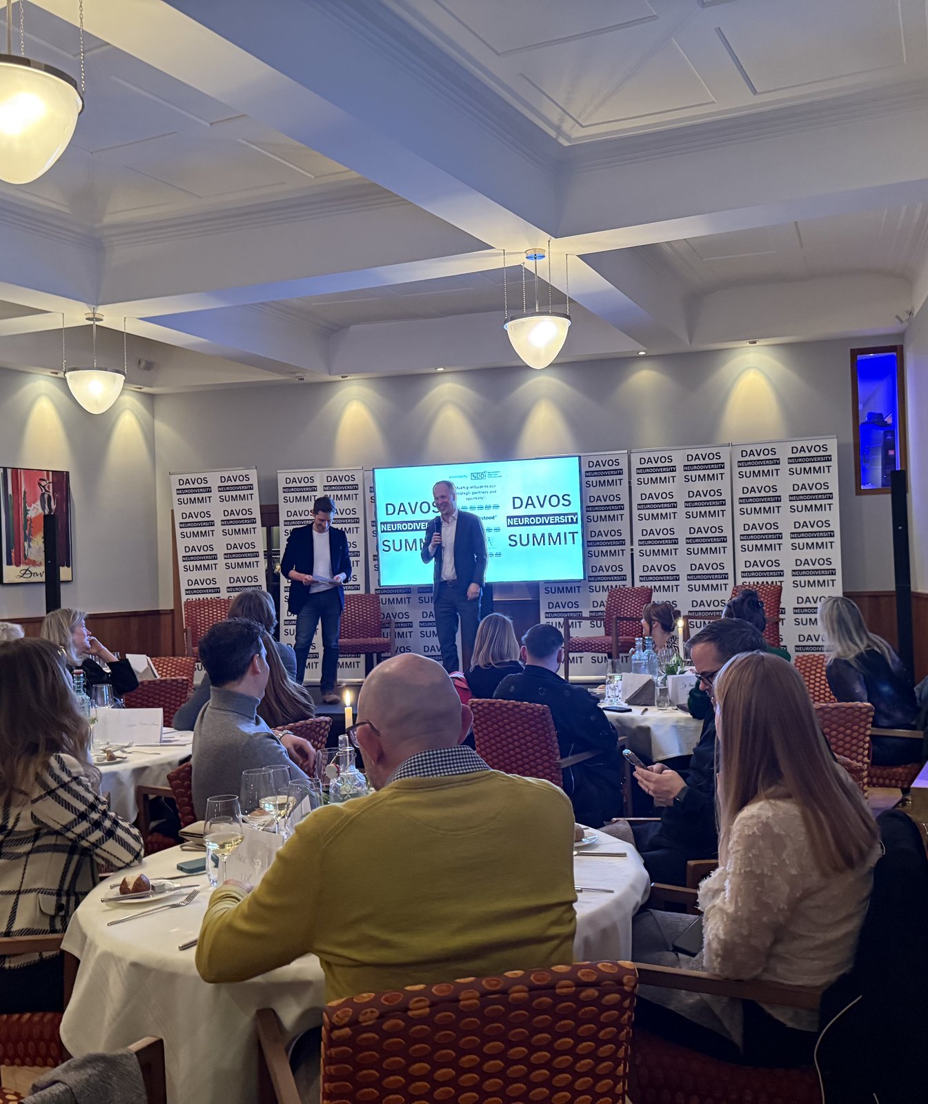 A speaker addressing the full dinner room at DNS, with Davos Neurodiversity Summit signage behind