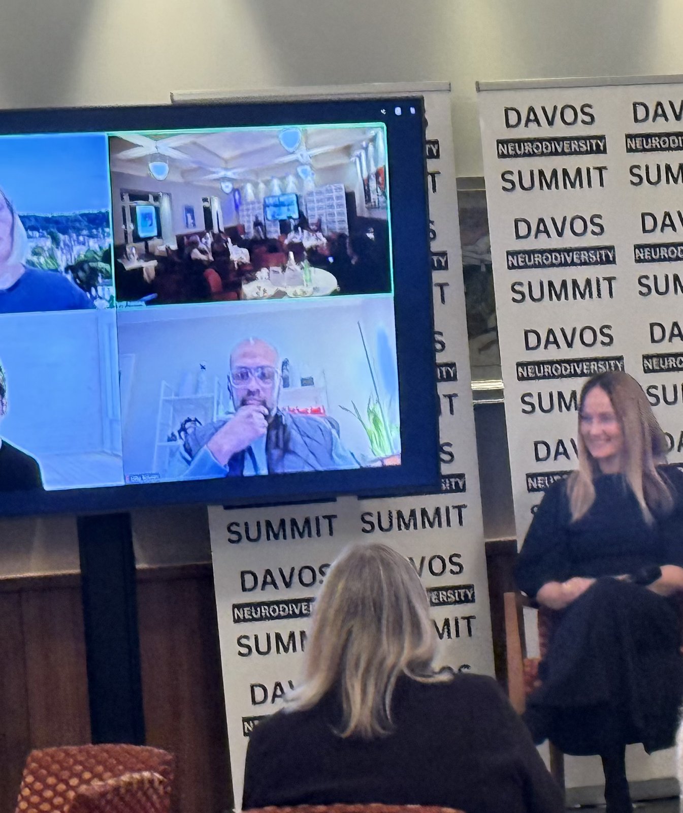 A hybrid panel — remote panelists on screen, with a moderator seated in the room